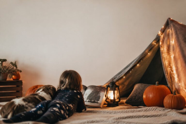 A child and a dog reading a book together in a lit-up indoor tent surrounded by pumpkins.